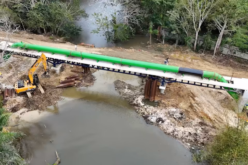 Vista aérea de la instalación de una gran tubería verde sobre una estructura metálica que cruza un río, con maquinaria de construcción y trabajadores